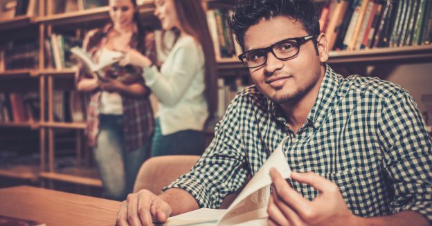 Male, college-aged student sitting in library with book and two female students in background